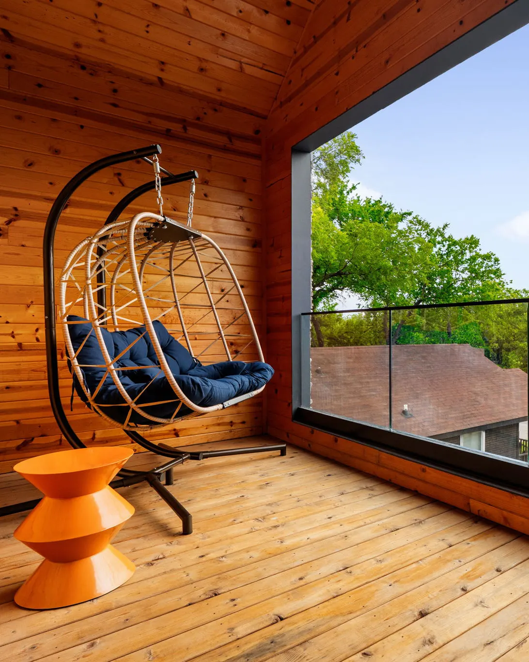 A cedar-lined balcony off the primary bedroom, with a hanging egg chair, an orange side table and a glass railing looking out over the treetops