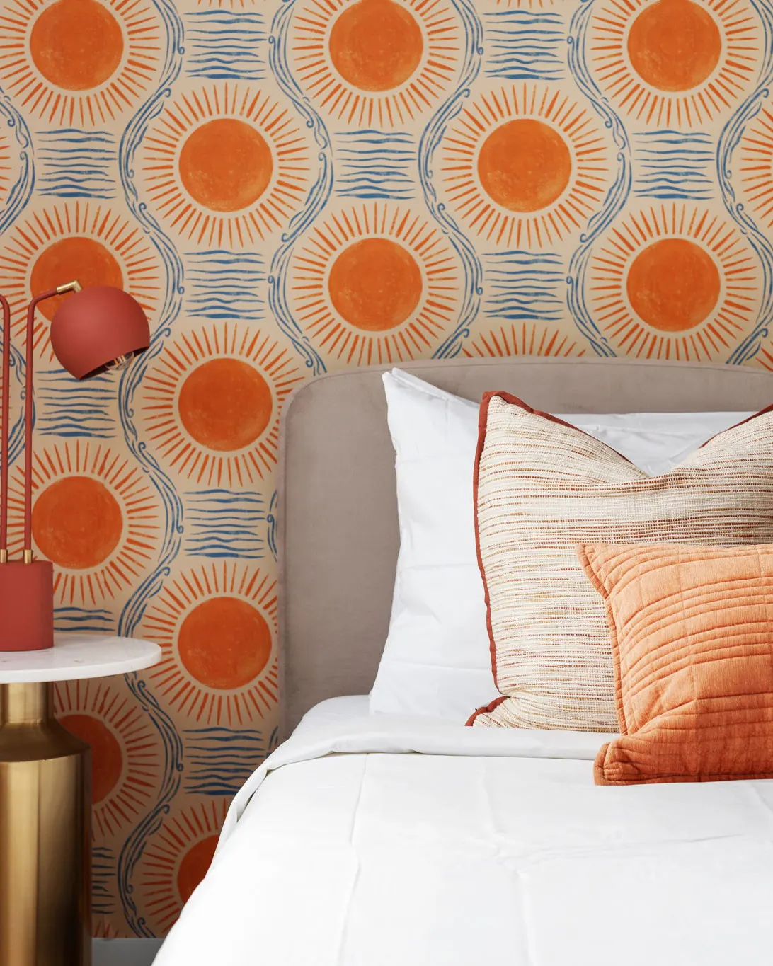 A bed with orange textured pillows against wallpaper printed with orange suns and blue waves, beside a red lamp