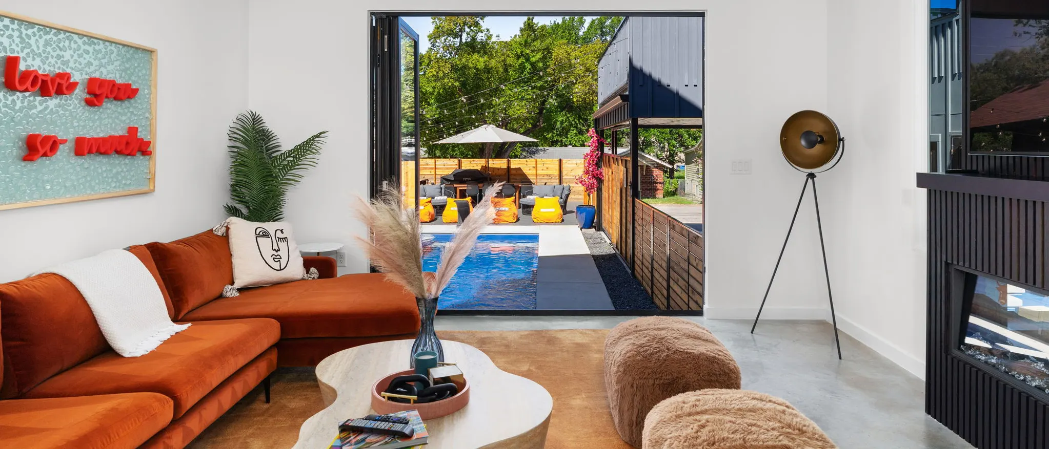 A white living room with a rust velvet sectional and a "love you so much" artwork, its glass doors folded open onto the pool and orange loungers
