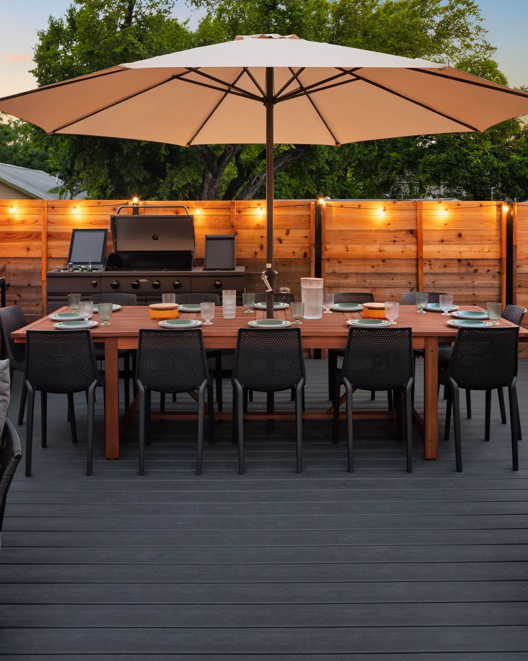 An outdoor dining table set for twelve under a large umbrella, with a grill and prep station against the fence