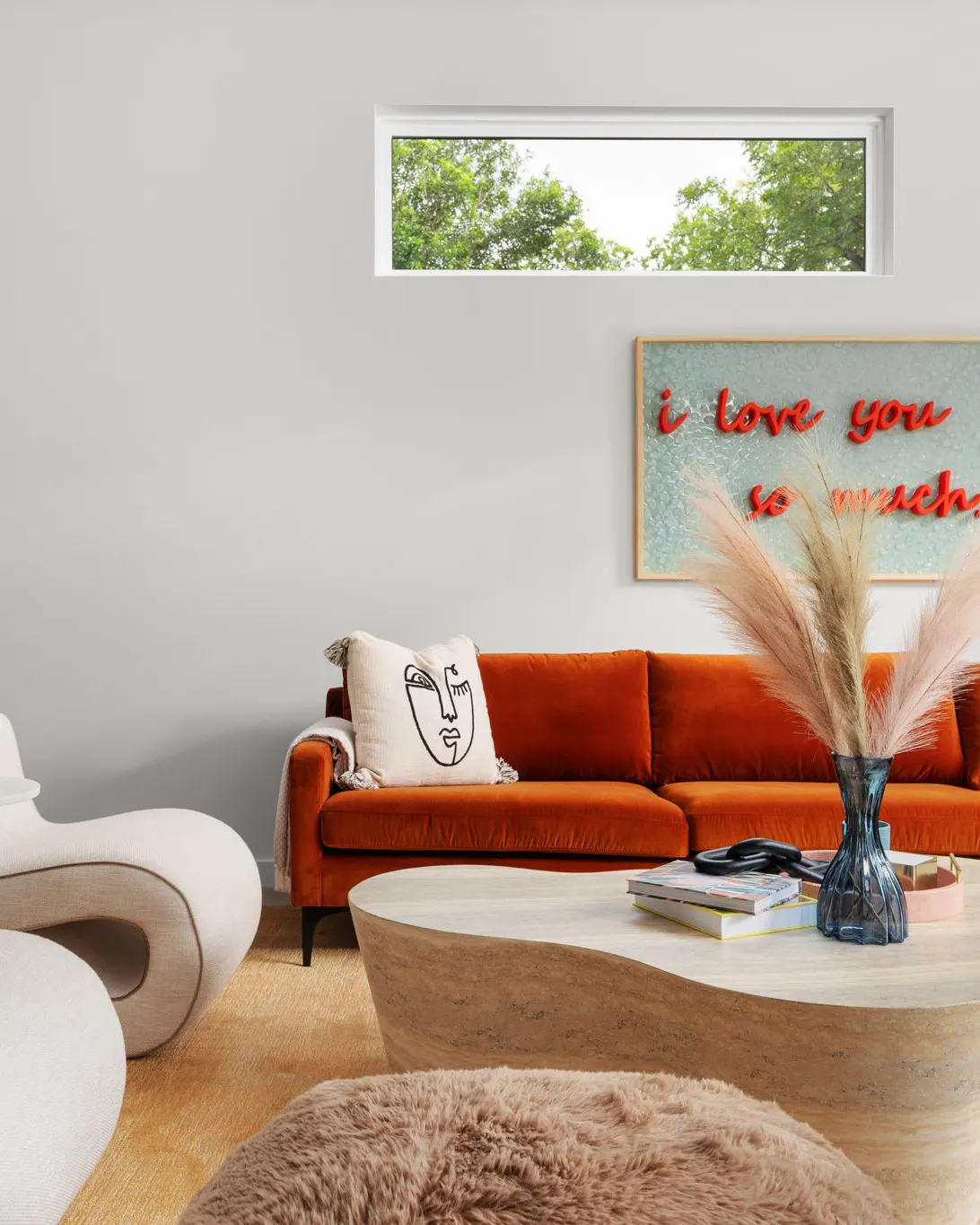 A rust velvet sofa beneath a "love you so much" artwork, with a travertine coffee table, pampas grass and a sculptural cream chair