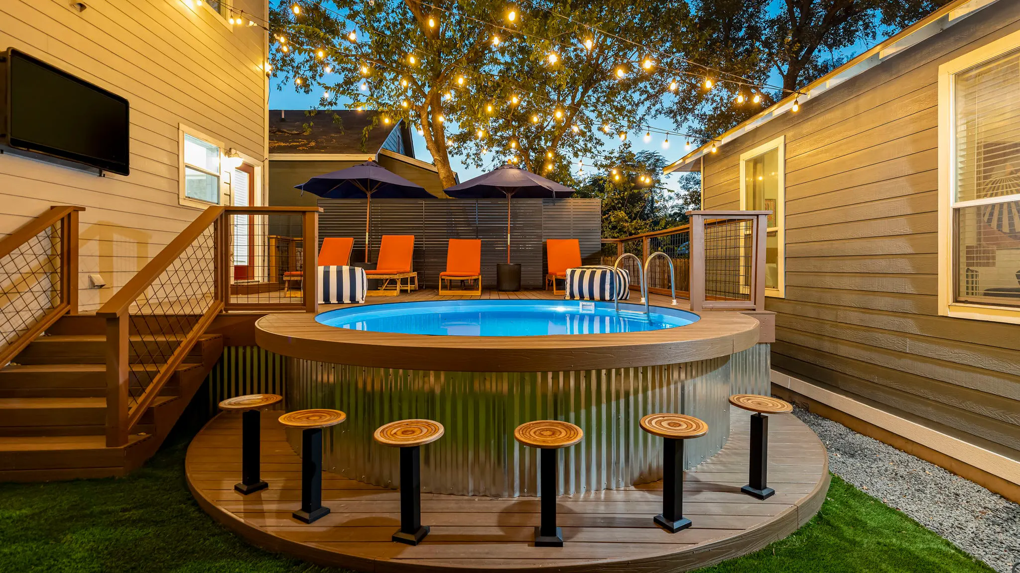 The above-ground pool at dusk with a deck built around it, bar stools along its edge, orange loungers under umbrellas and a TV mounted on the house