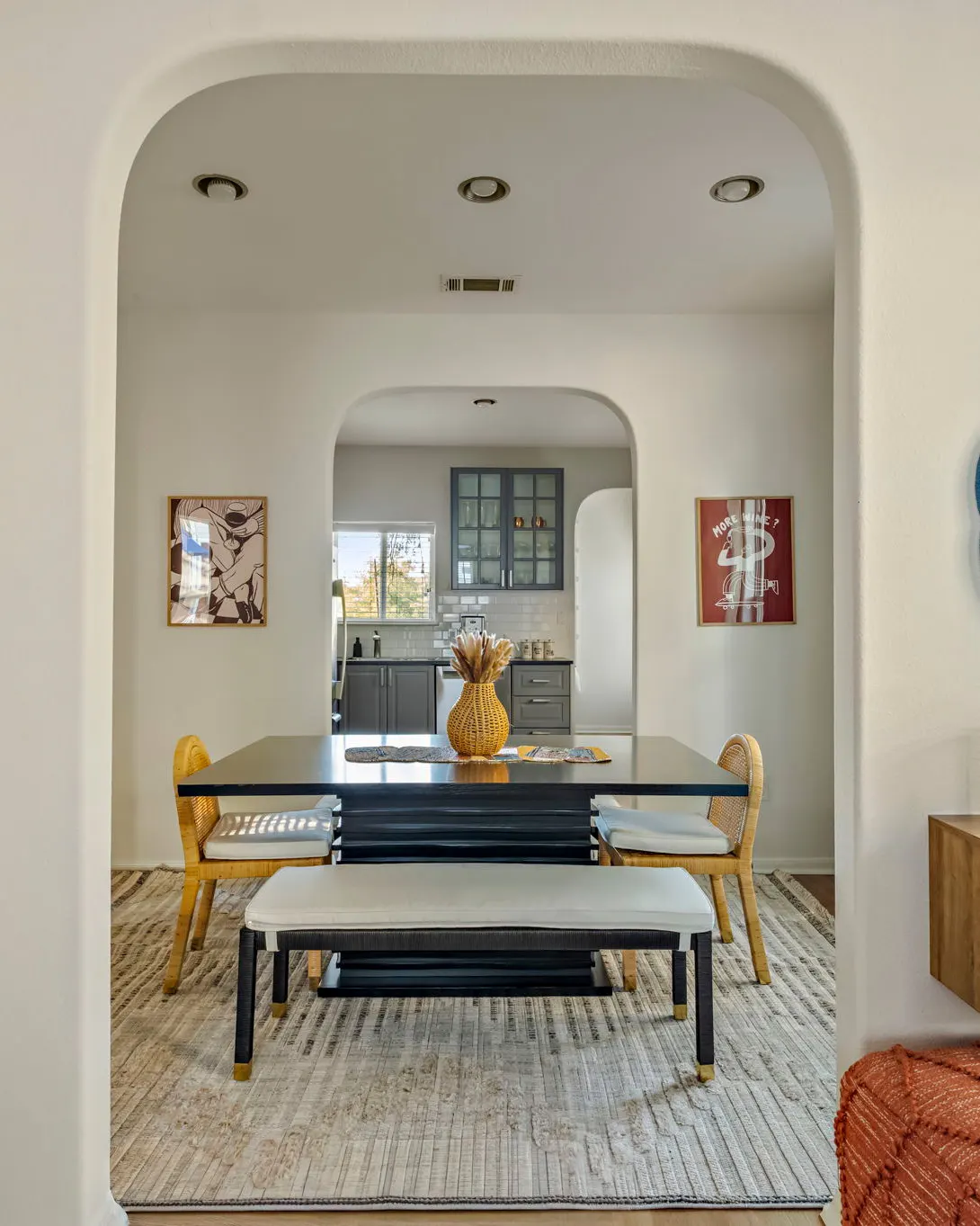An arched doorway framing a dining table with rattan chairs and a bench, with a grey kitchen through a second arch