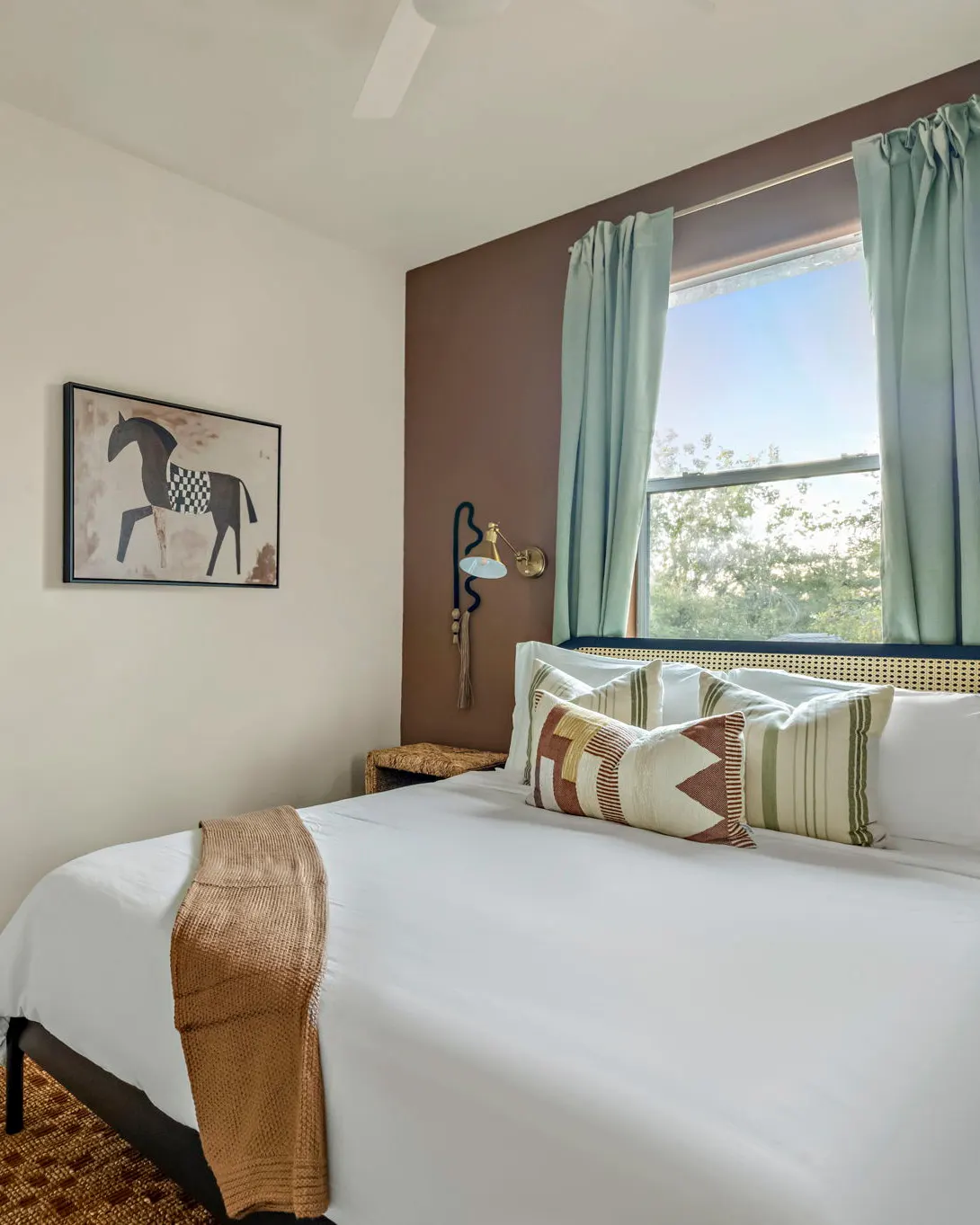 A bedroom with a brown accent wall, a cane headboard, a wavy wall sconce and a horse print