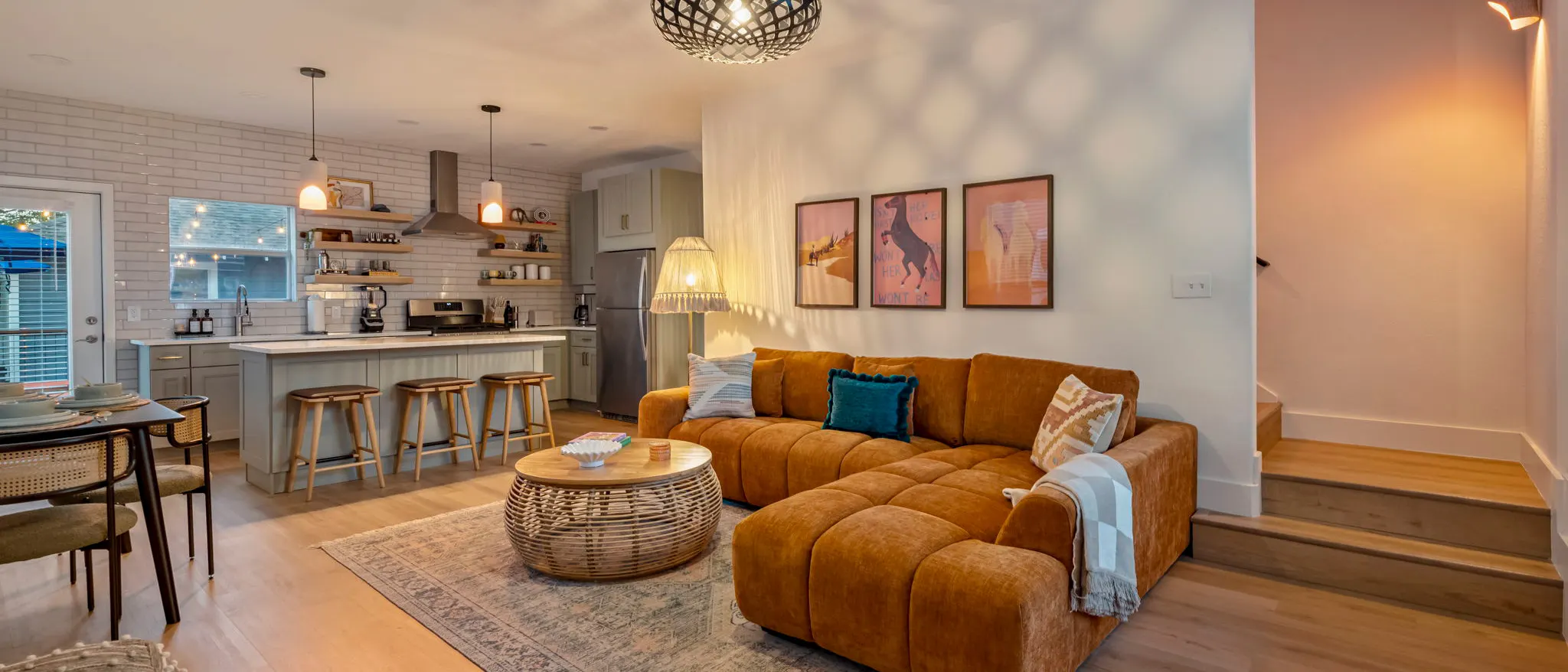 An open-plan living room with a caramel velvet sectional beneath three desert and horse prints, a rattan coffee table, and the kitchen island with wooden stools behind
