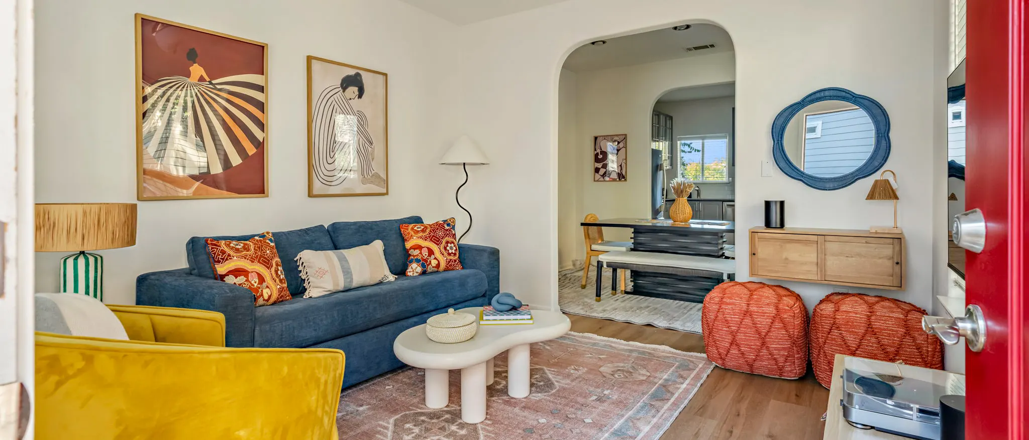 A living room seen from the red front door: a blue sofa beneath two fashion prints, a mustard velvet armchair, orange poufs, and an arch through to the dining table