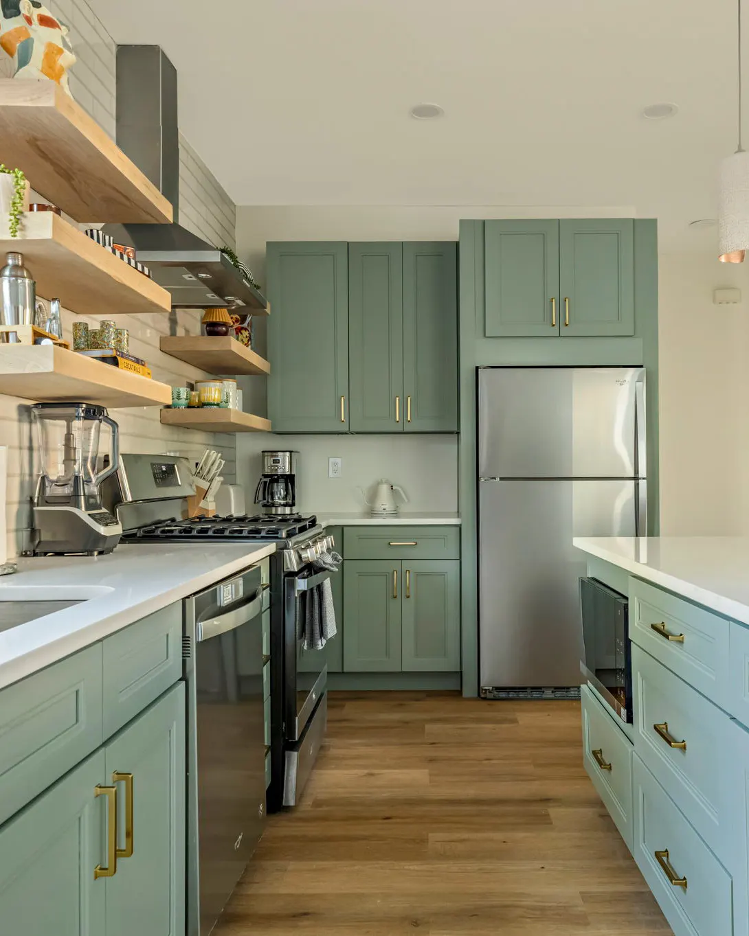 A kitchen with sage green cabinets and brass pulls, open wooden shelves, and the end of the island in the foreground