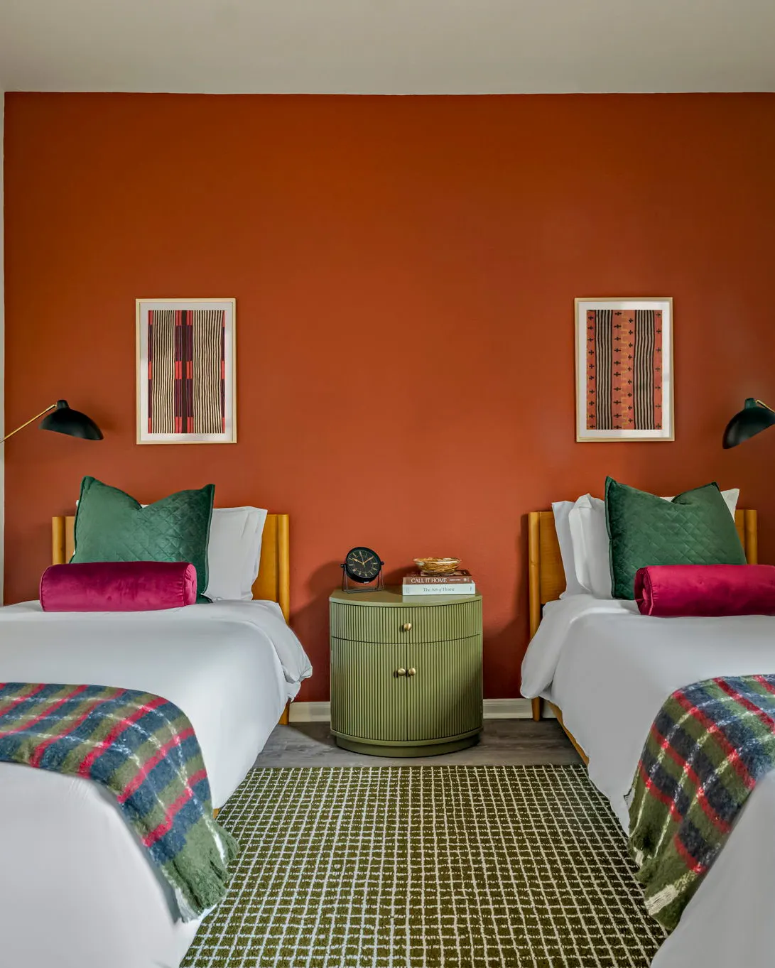 Two twin beds with green quilted pillows and plaid throws against a terracotta accent wall, with a ribbed green nightstand between them