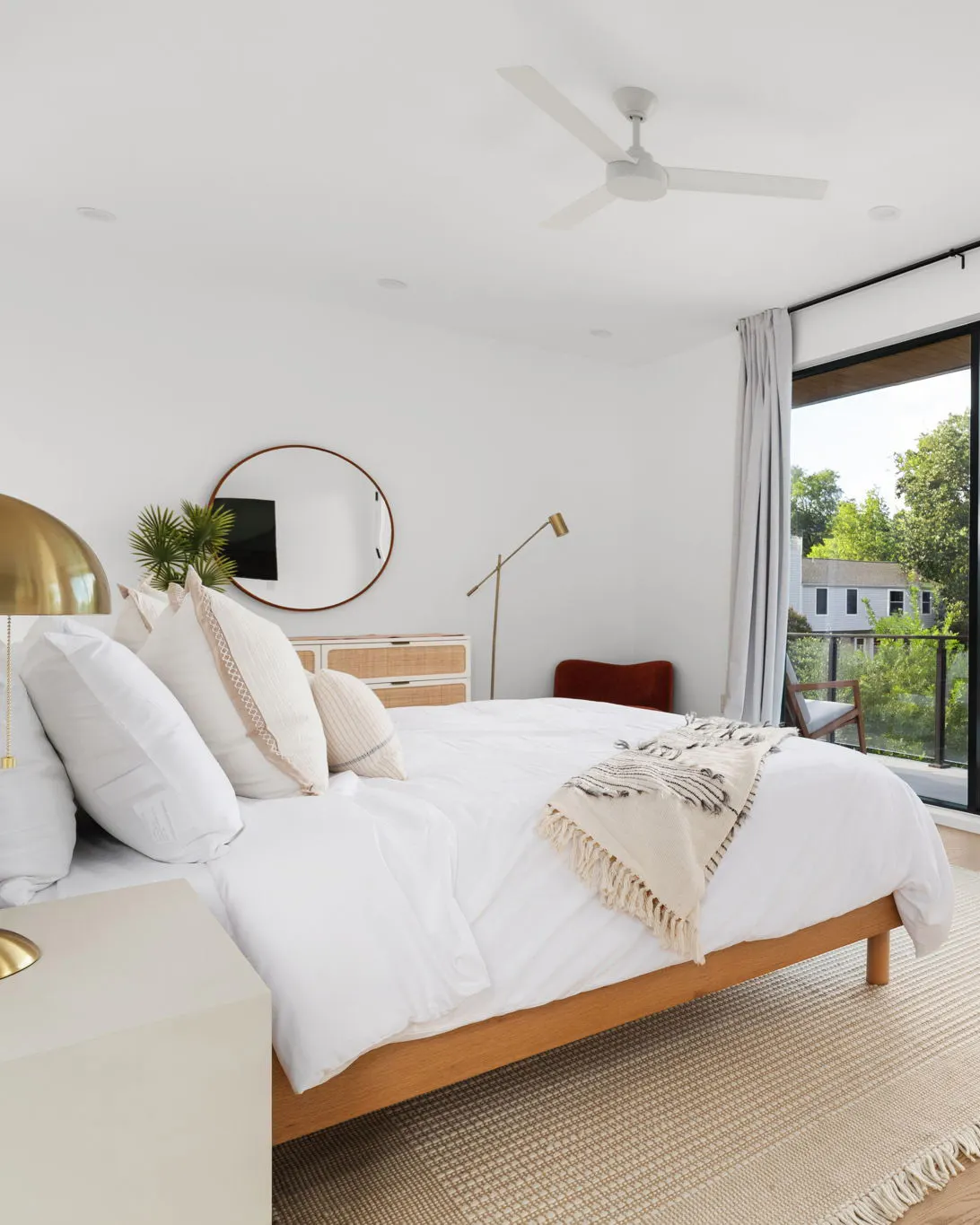 A bedroom with a white bed on an oak frame, a round mirror, a brass floor lamp and a glass door onto a balcony