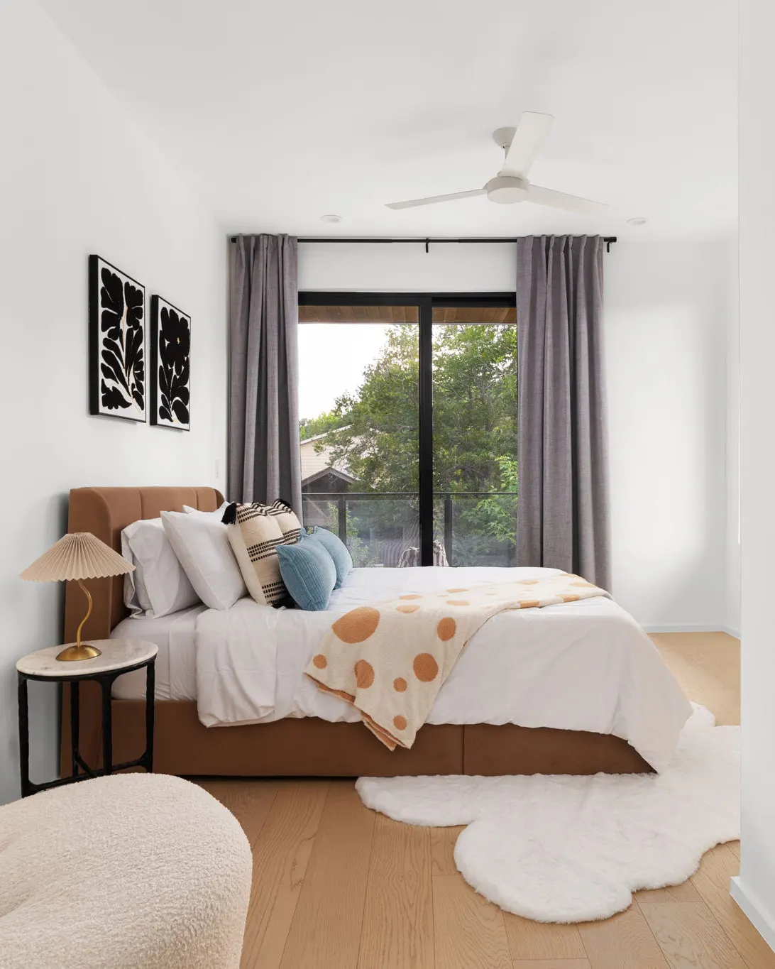 A bedroom with a tan upholstered bed, a polka dot throw, a cloud-shaped sheepskin rug and a glass door onto a balcony