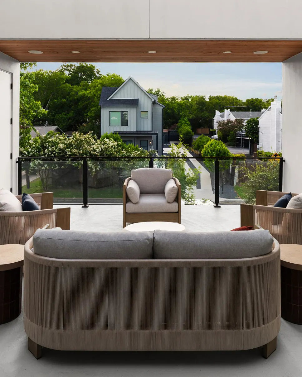 A covered balcony with a curved rope sofa and teak armchairs, looking out over the treetops and the street