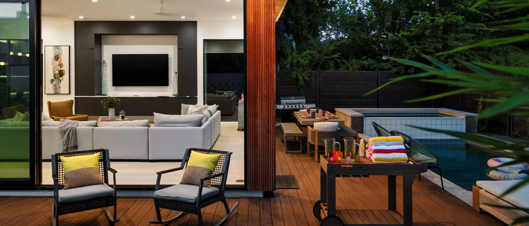 The deck at dusk: two rocking chairs with yellow pillows and a bar cart stacked with striped towels, beside the living room, the outdoor dining table and the spa