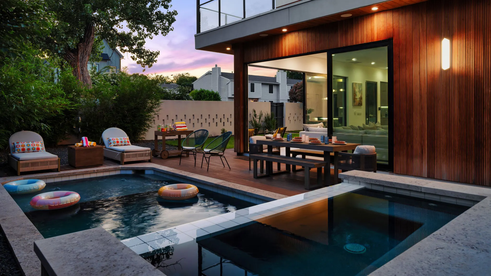 The pool and spa at dusk with inflatable rings, two loungers with patterned cushions, a bar cart, and an outdoor dining table beside the house