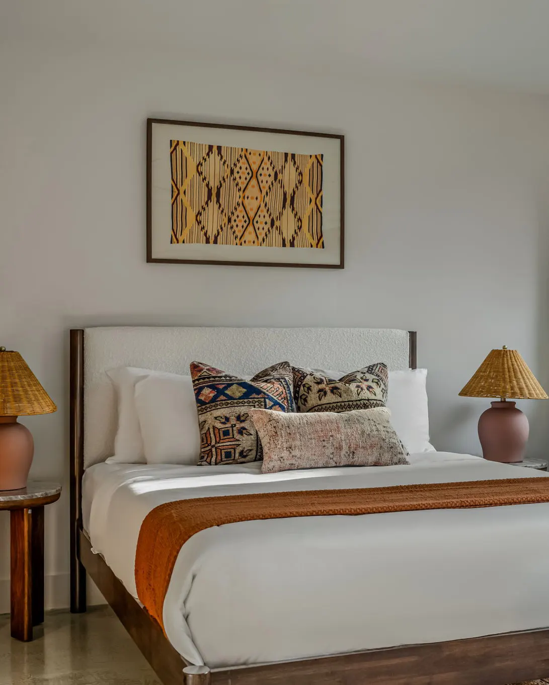 A bed with a cream bouclé headboard, patterned pillows and a rust throw, between two terracotta lamps with woven shades