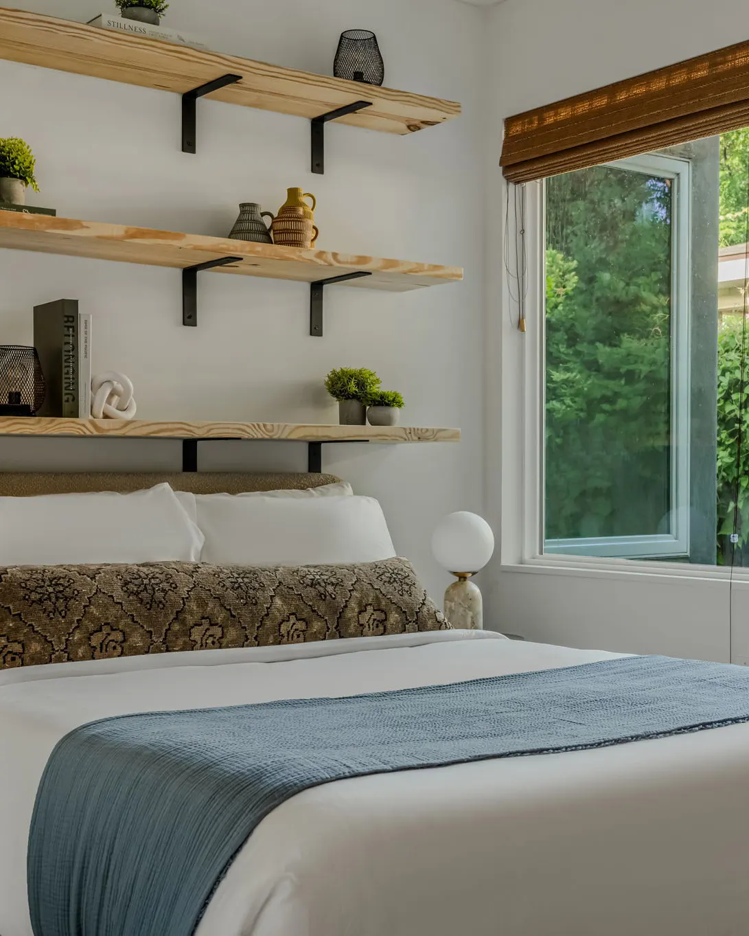 A bed with a patterned bolster and a blue throw beneath three floating wooden shelves, beside a window onto green trees