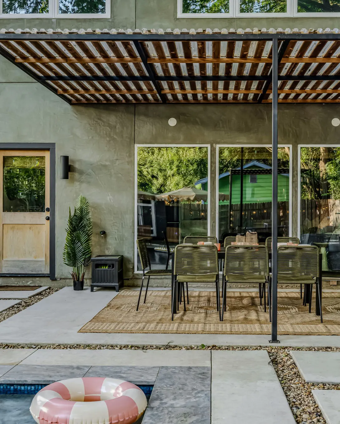 An outdoor dining table with green rope chairs under a pergola, seen from the edge of the pool