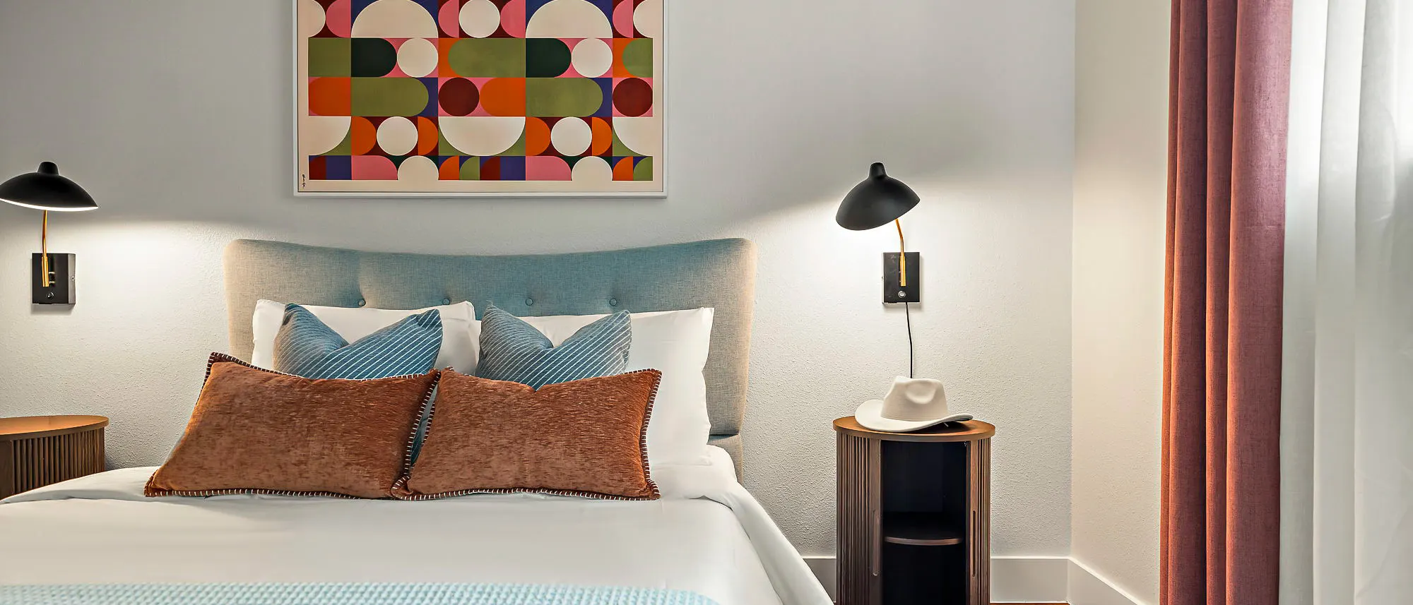 A bed with rust pillows and a blue tufted headboard beneath a geometric print, between two black wall lamps, with a white cowboy hat on the nightstand