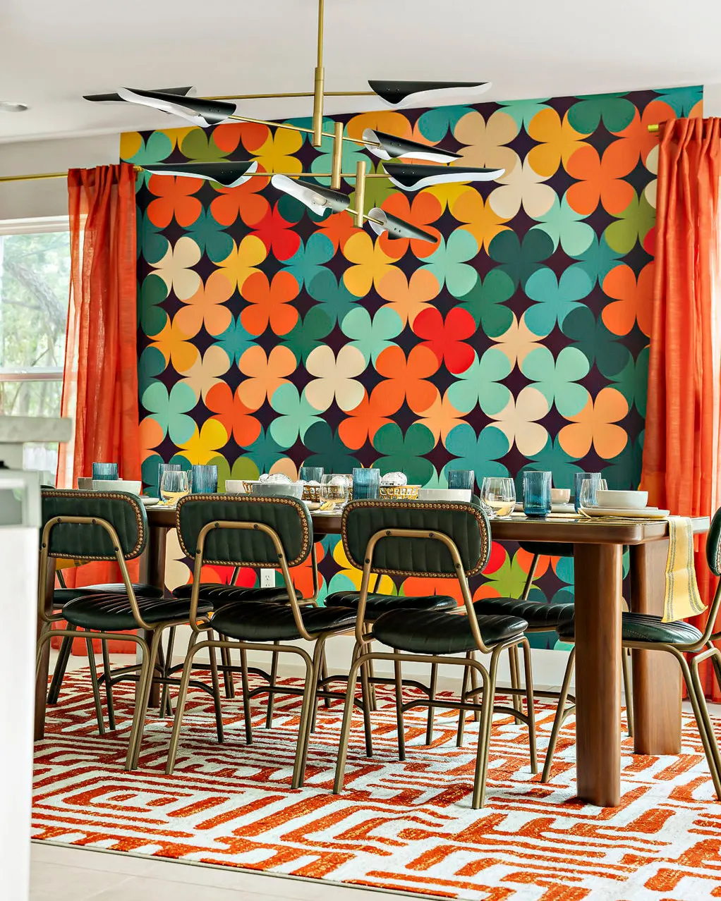 A dining table with green leather chairs on an orange patterned rug, beneath a sculptural brass chandelier, against a wall of bold orange, teal and cream quatrefoils