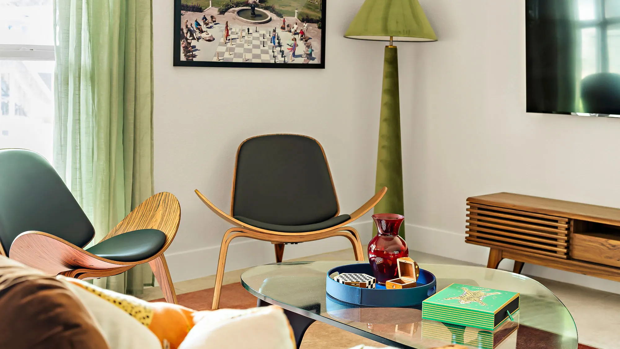 Two bentwood shell chairs beside a green velvet floor lamp and a glass coffee table, beneath a framed photograph of people around a giant chessboard