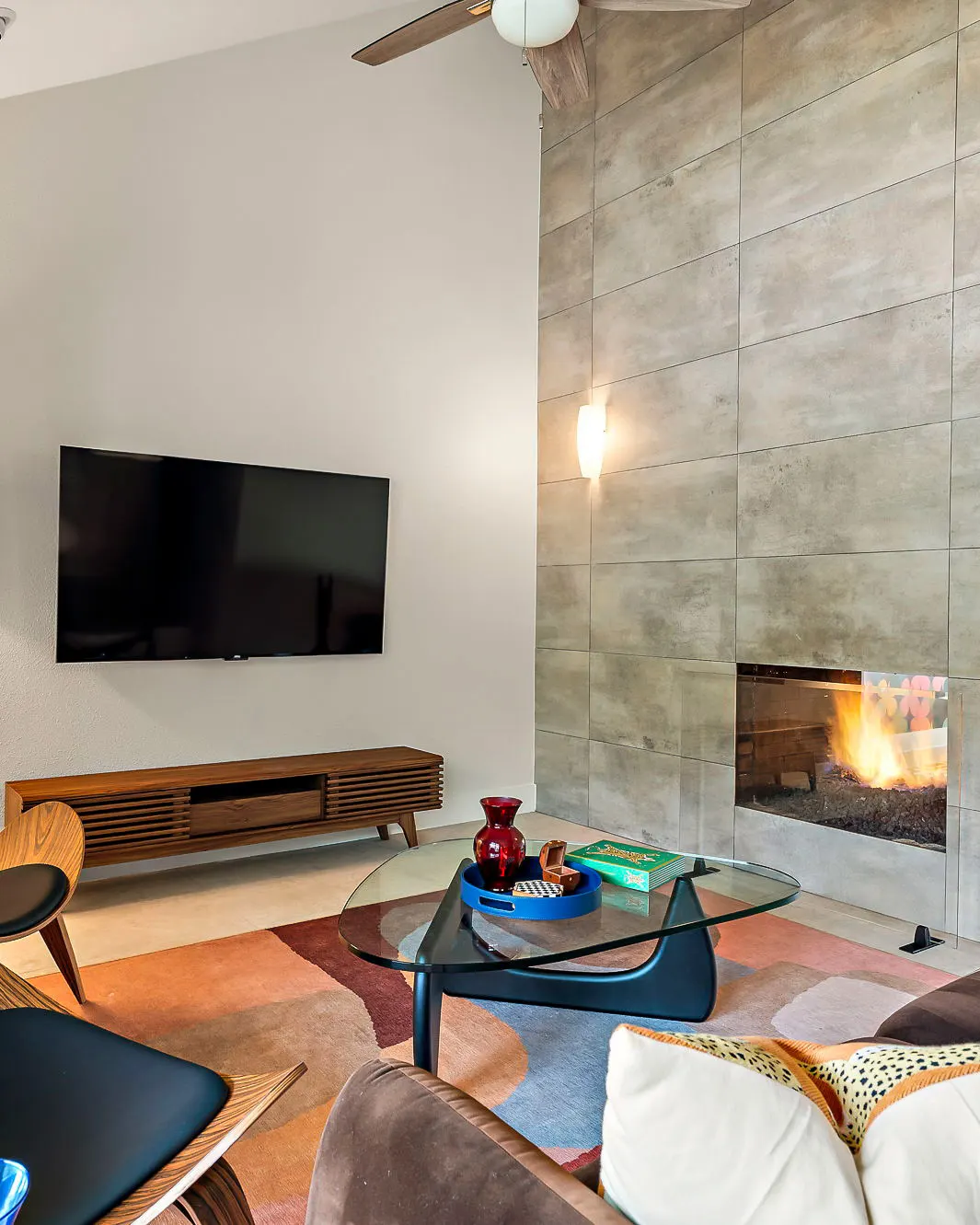 A living room with a tall tiled fireplace, a mid-century media console and TV, and a glass coffee table on a colorful rug