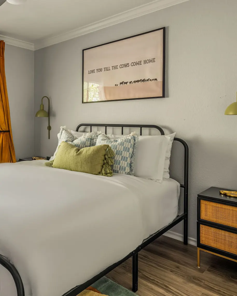 A bedroom with a black iron bed, green and blue pillows, and a "Love you till the cows come home" print