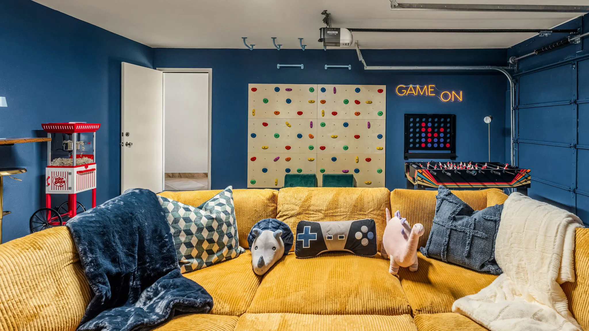 The mustard corduroy sectional with animal and game controller pillows, facing the climbing wall, the "Game On" neon, a giant Connect Four, foosball and a popcorn machine