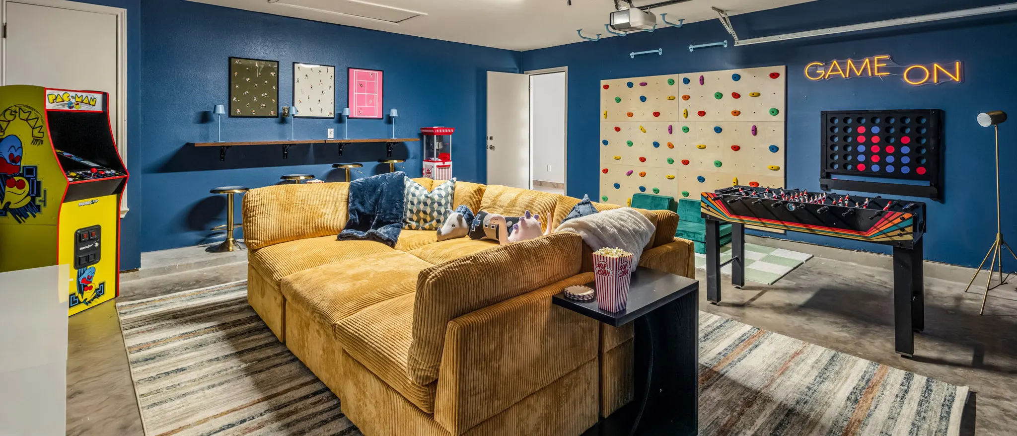 The garage game room: a mustard corduroy sectional facing a climbing wall, a Pac-Man arcade machine, foosball, a giant Connect Four and a neon "Game On" sign on navy walls