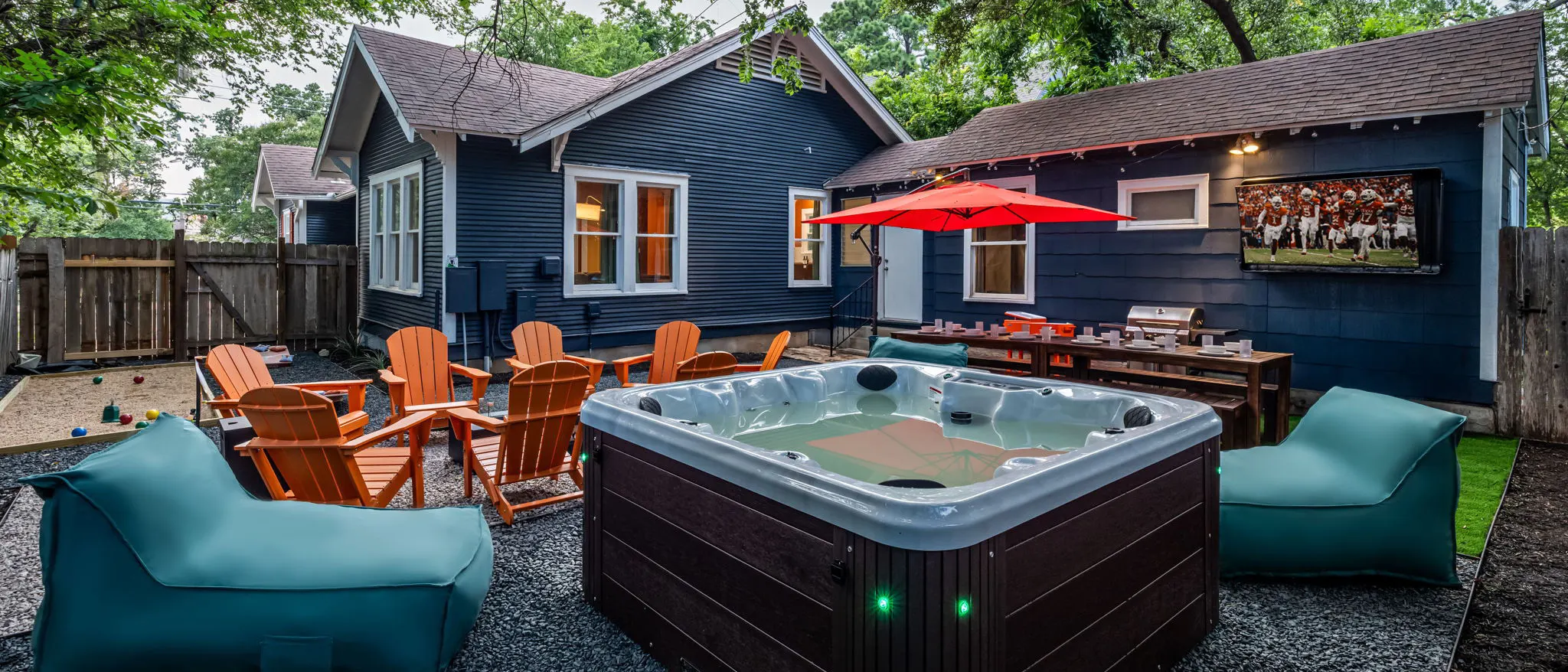 The backyard: a hot tub with teal lounge chairs, orange Adirondack chairs, a bocce court, and a dining table under a red umbrella beside an outdoor TV showing a football game