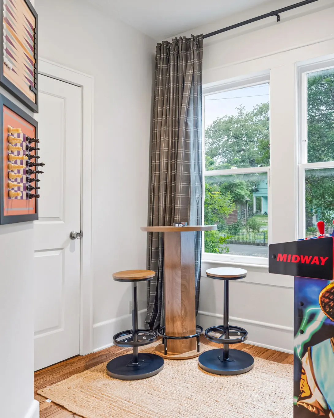 A corner with a high-top table and two stools by the windows, framed game boards on the wall, and a Midway arcade machine