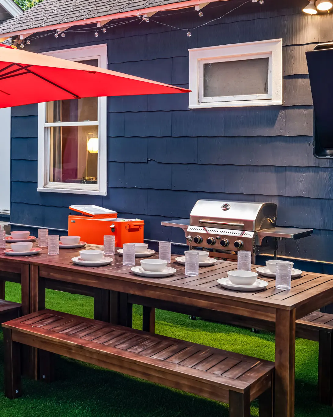 An outdoor dining table set with bowls and glasses, with an orange cooler, a grill and a mounted TV against the navy house