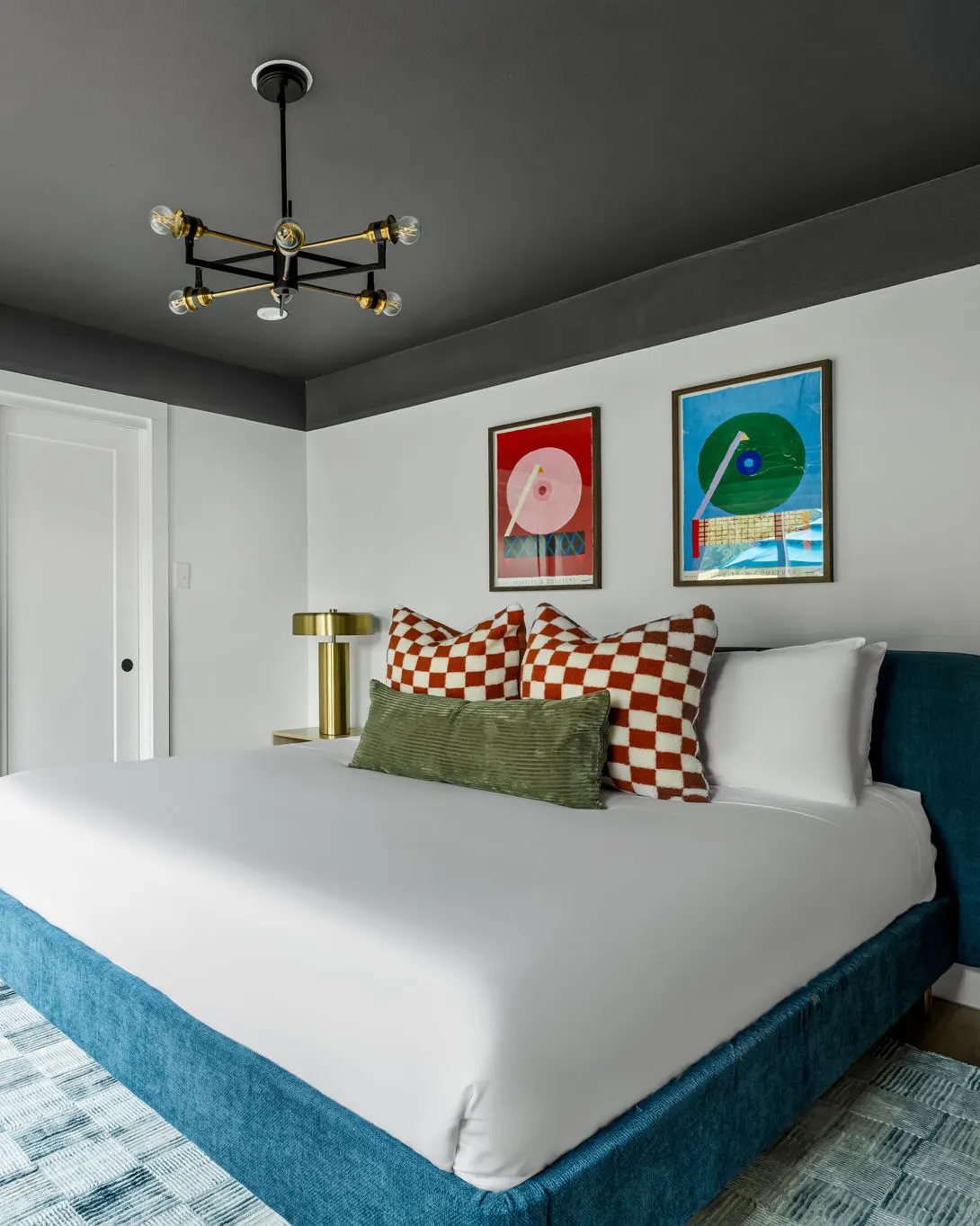 A bedroom with a teal upholstered bed, checkered pillows, a charcoal ceiling and two colorful turntable prints