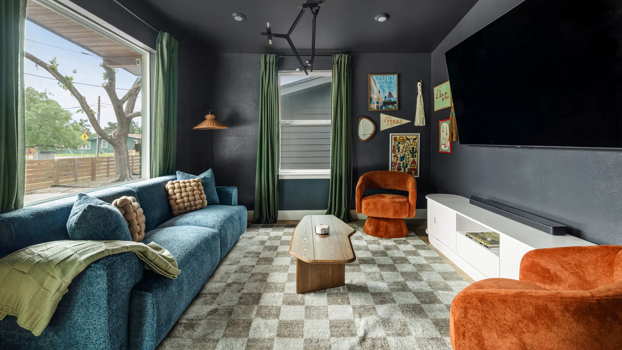 A charcoal media room with a blue bouclé sofa, orange armchairs, a checkered rug, a large TV and a wall of Austin and Texas artwork